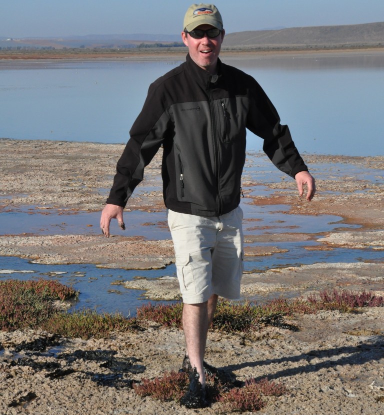 Richard Craig Everroad at what appears to be a marshland wearing sunglasses, a tan hat, black jacket, and