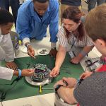 A group of students stand work on small payload in the center of a table.