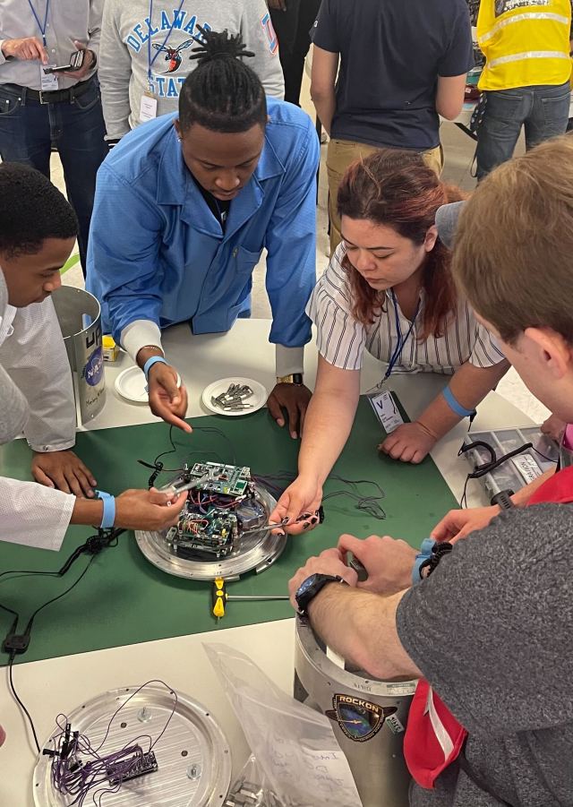 A group of students stand work on small payload in the center of a table.