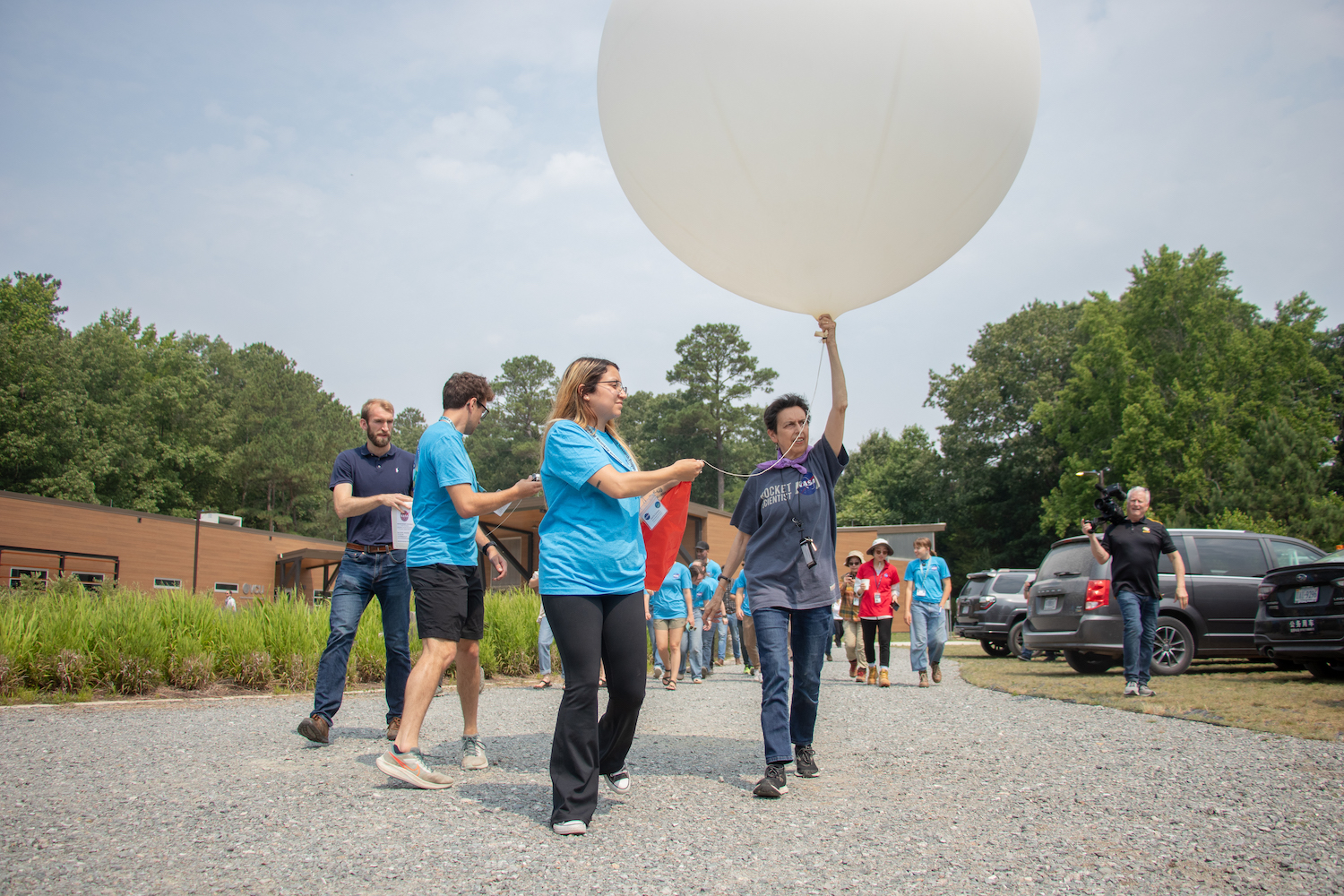 A group of students and advisors walk along a concrete path, a cloudy blue sky and green trees in the background. An advisor, centered in the image, is holding a very large, white, spherical balloon above her head.