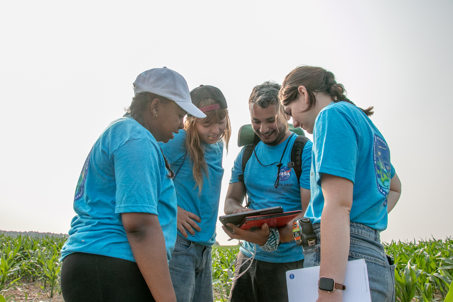 Four people stand in a corn field. They're wearing matching blue t-shirts, with the NASA logo on the front and a SARP mission logo on the back. Th