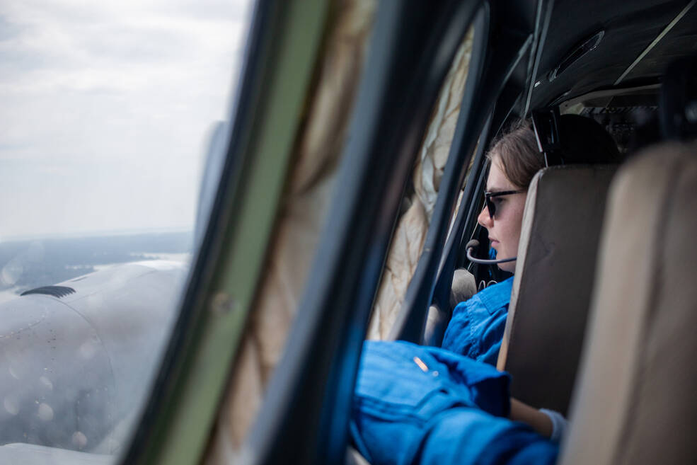 A student in a headset on looks out a window of a plane. The rim of another window cuts down the middle of the image separating the interior and exterior of the plane. The engine and propellors are visible through the window.