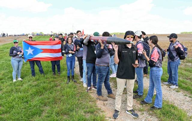 Students from the University of Puerto Rico, Mayaguez Campus, carry their rocket and flag on a grassy field.