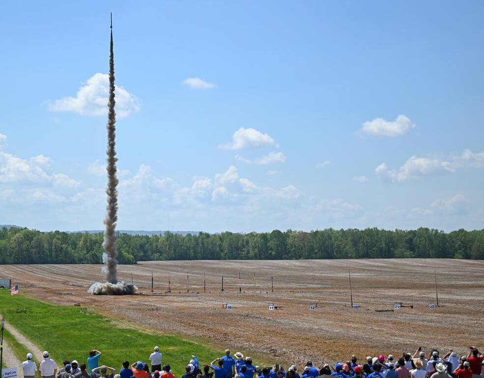 A student rocket launches into the blue sky from a field.