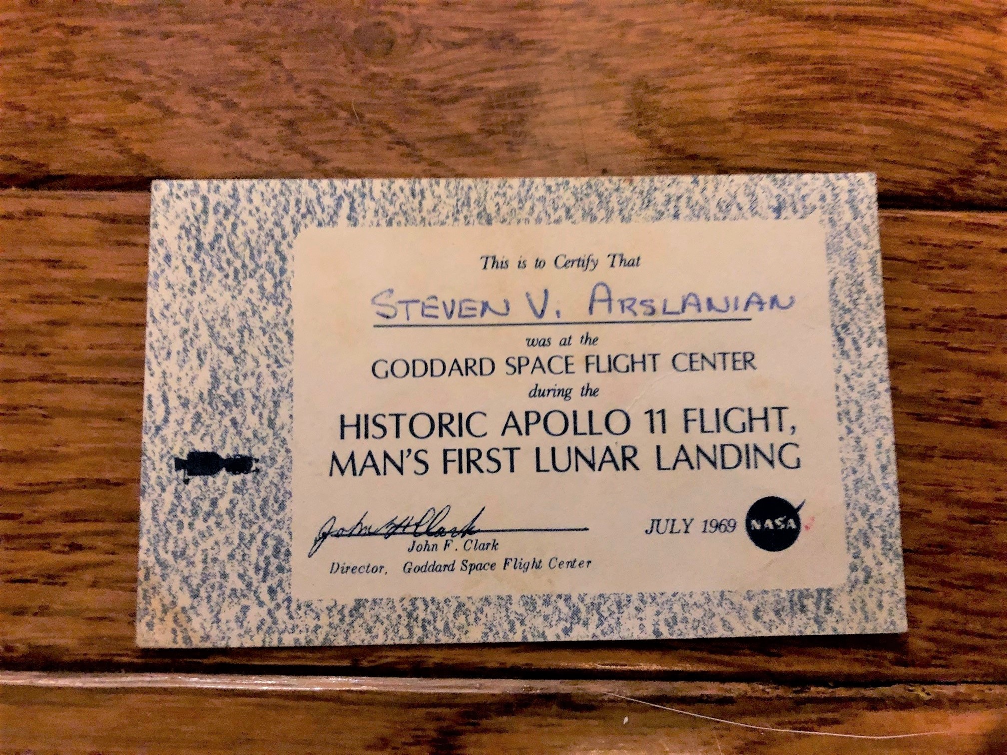 A card sitting on a table. Text reads "This is to certify that Steven V. Arslanian was at the Goddard Space Flight Center during the historic Apollo 11 flight, man's first lunar landing." A signature, date (July 1969), and logo are at the bottom.