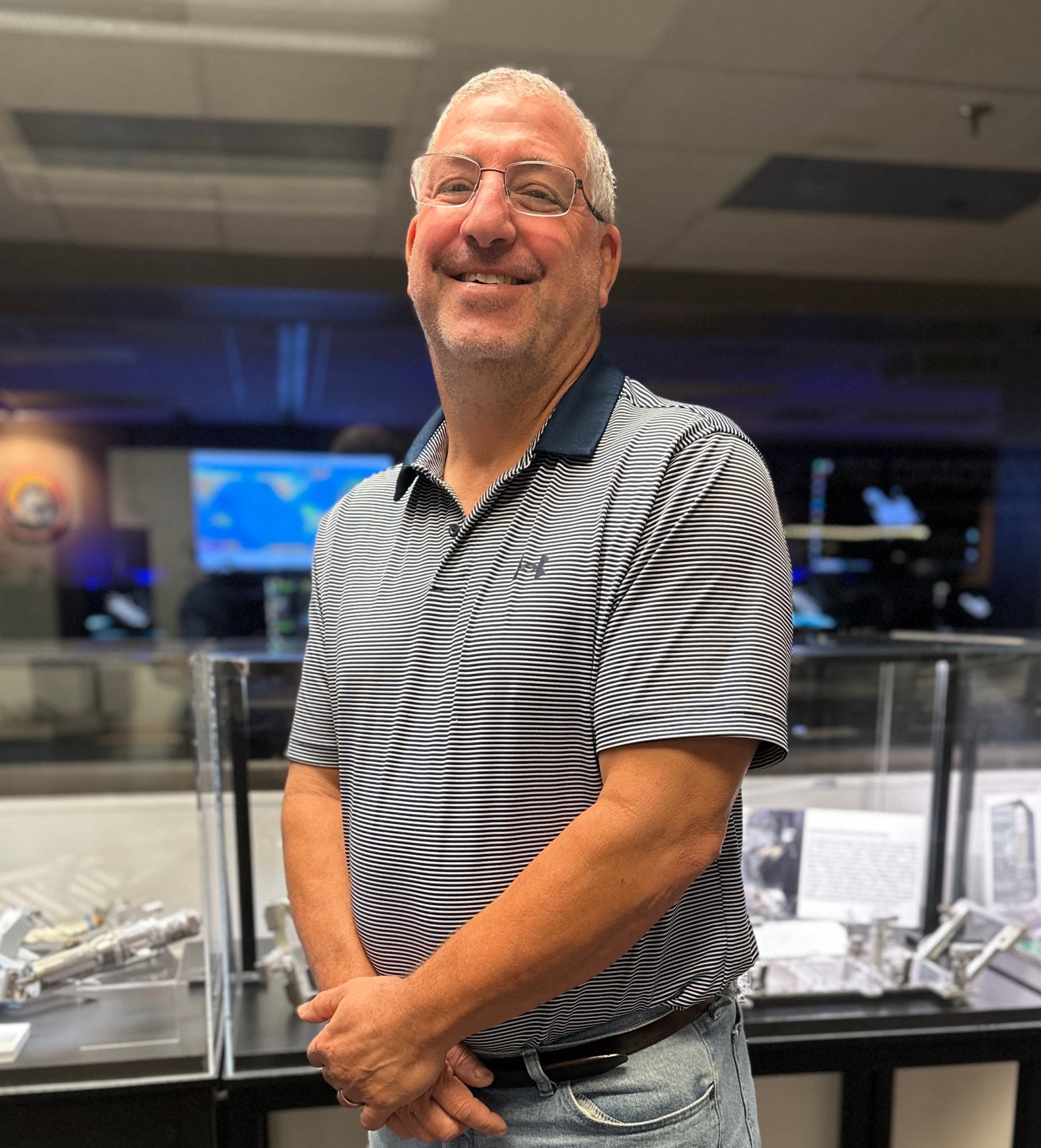 Steve Arslanian, a man with white hair and glasses, smiles and stands with hands folded in front of him, wearing a navy striped polo shirt and faded jeans. He stands in front of display cases and the Hubble control room with monitors in the background.