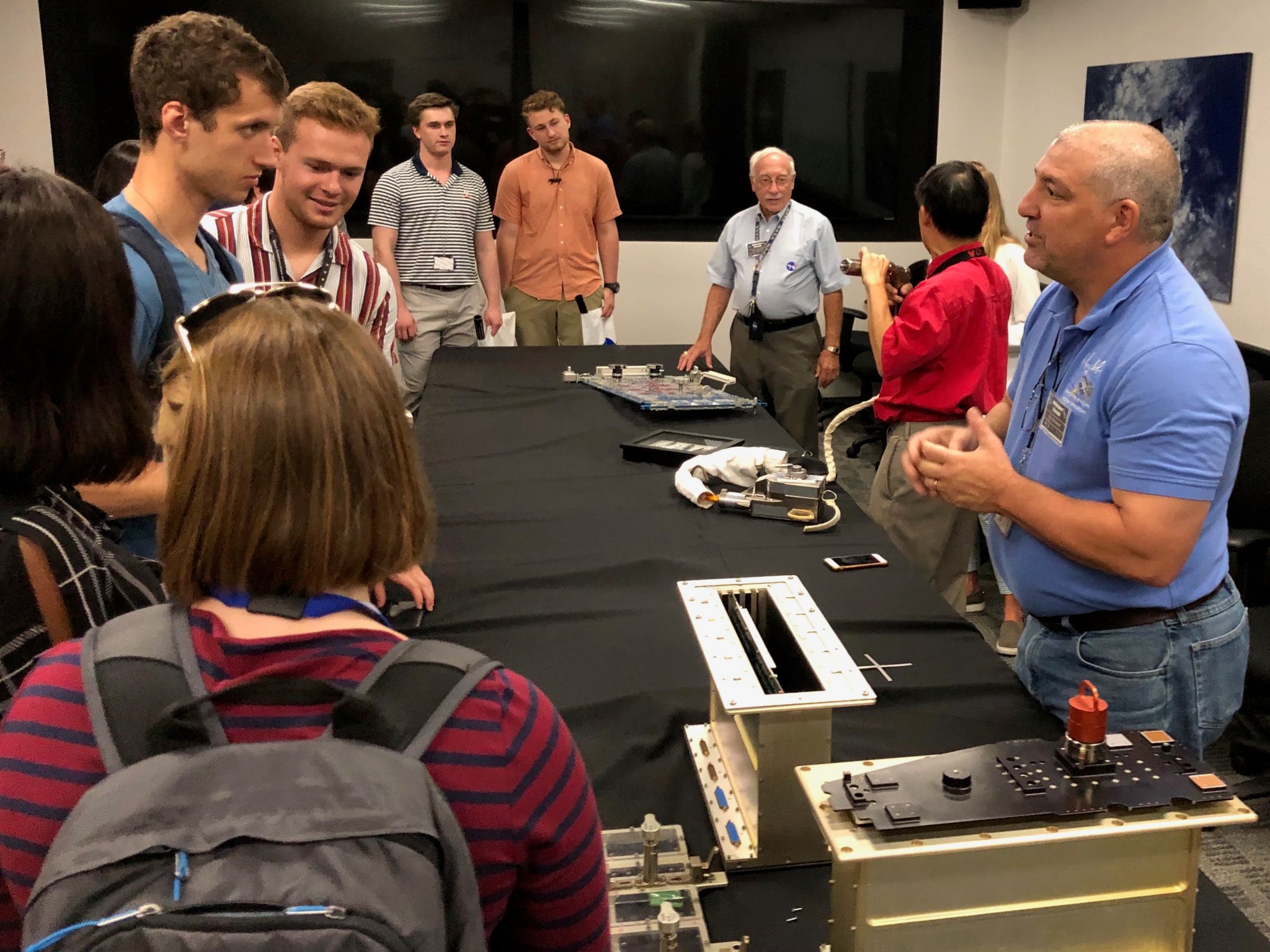 Steve Arslanian, a man with short gray hair, stands behind a table speaking to a class of students. He wears a blue polo with a logo and jeans. The table has various electronics and technology on top of a black tablecloth.