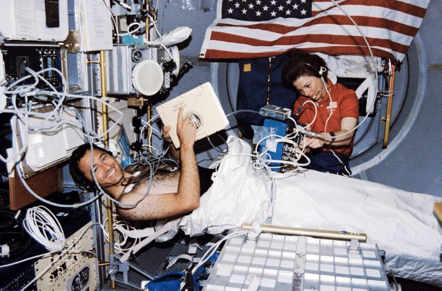Mission Specialist Bonnie Dunbar and Payload Specialist Lawrence DeLucas in the spacelab with the Lower Body Negative Pressure Study. DeLucas is encased in the suit and Dunbar is administering and overseeing the procedure