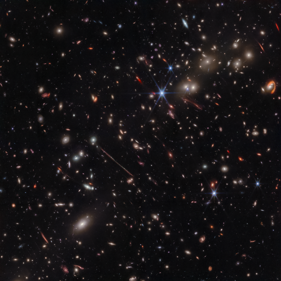 A black background is scattered with hundreds of small galaxies of different shapes, ranging in color from white to yellow to red. Some galaxies are distorted, appearing to be stretched out or mirror imaged.