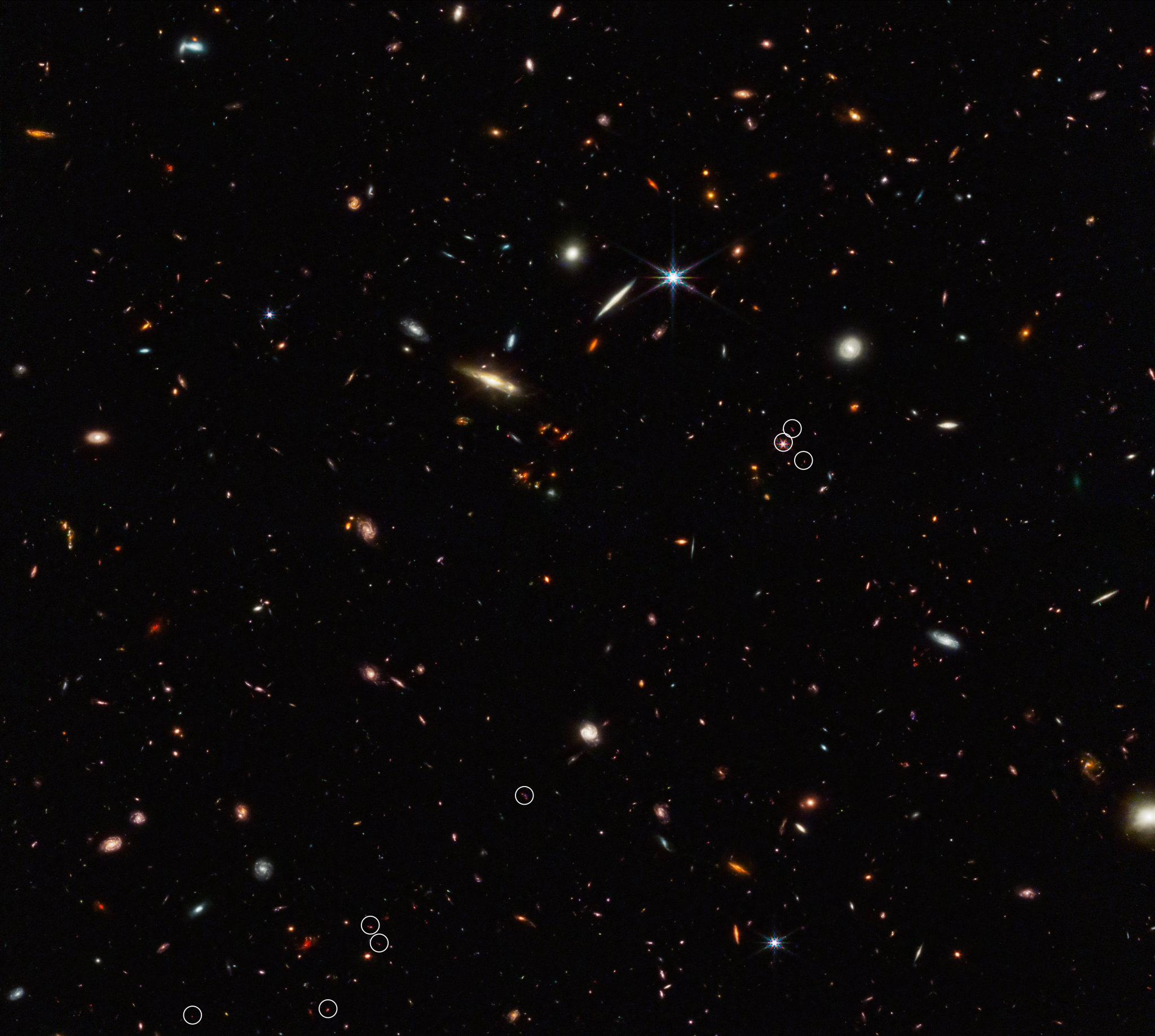 A deep field image with many colorful galaxies and six-pointed stars sprinkled across deep black space. Small white circles scattered across the center and lower left mark ten galaxies that existed just 830 million years after the Big Bang.