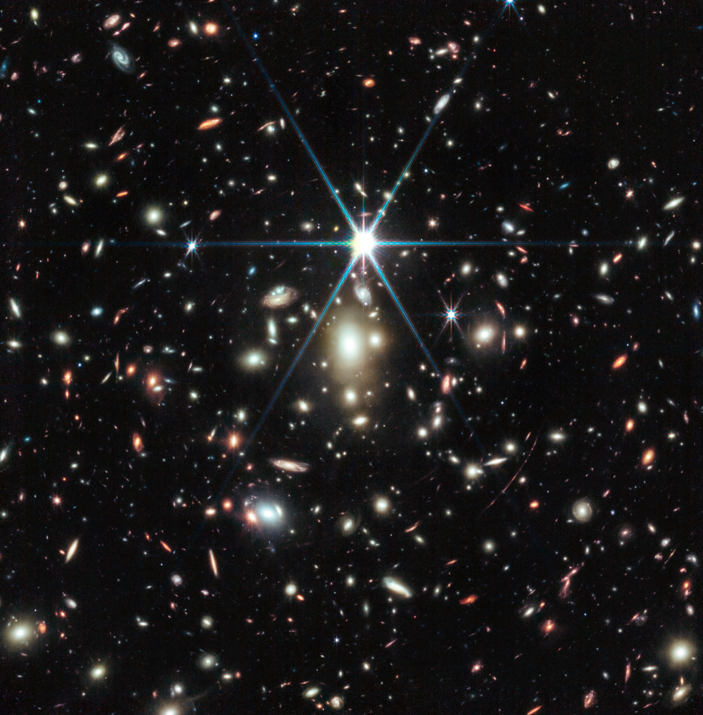 A black background is scattered with hundreds of small galaxies of different shapes, ranging in color from white to yellow to red. Just a bit above the center, there is a bright source of light, a star, with 8 bright diffraction spikes extending out.