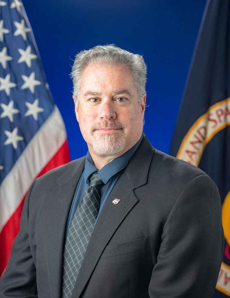 Studio portrait of Anthony Colaprete with the American and NASA flags in the background.