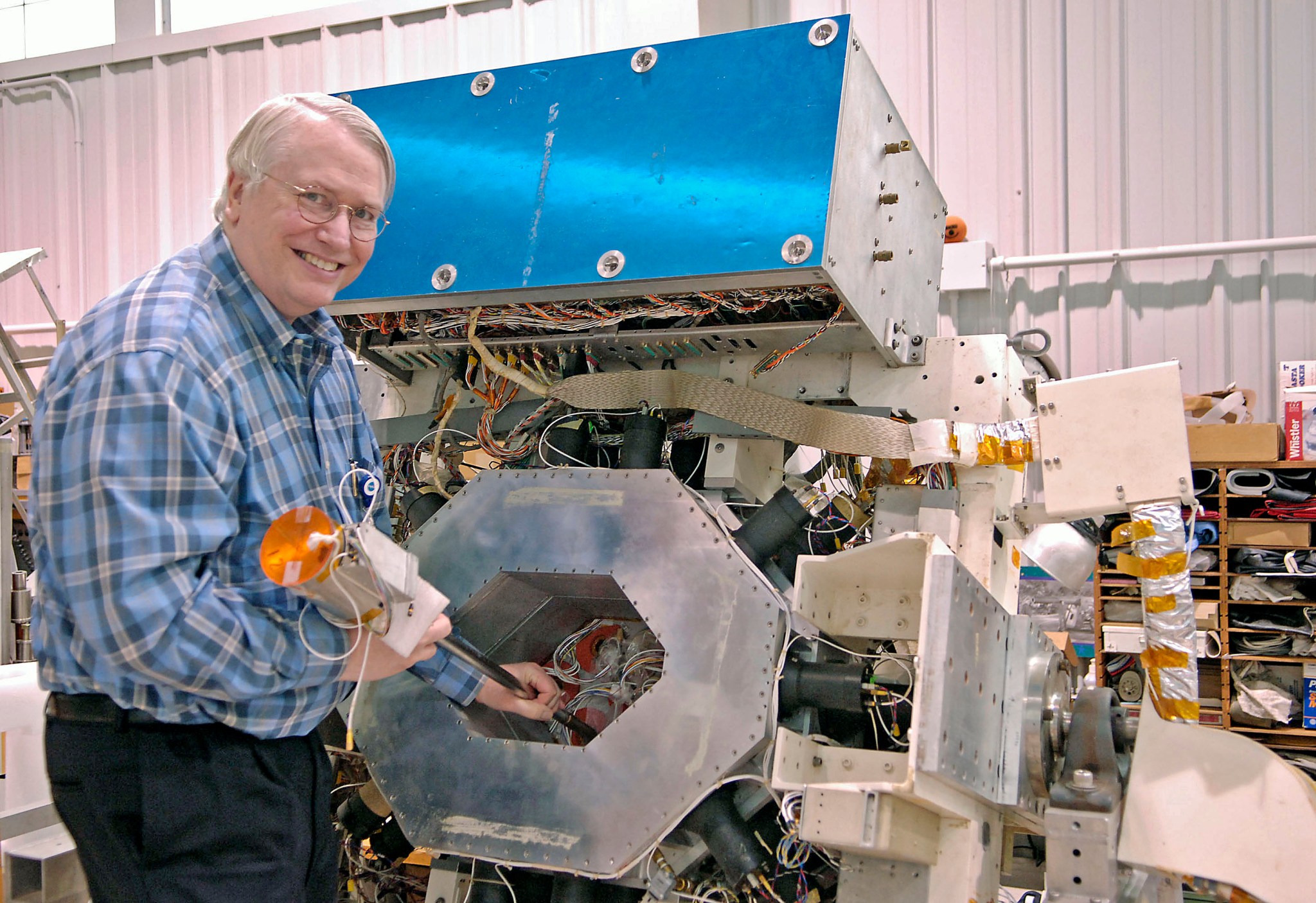 A man in a plaid shirt at left holds a cylindrical component near a large piece of steel-gray hardware. At top, one element is colored bright blue.