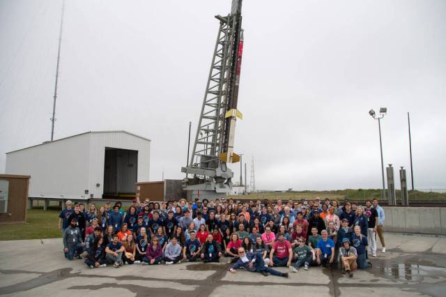A large group of people poses in front of a tall frame holding a small black, red, and yellow sounding rocket vertically.