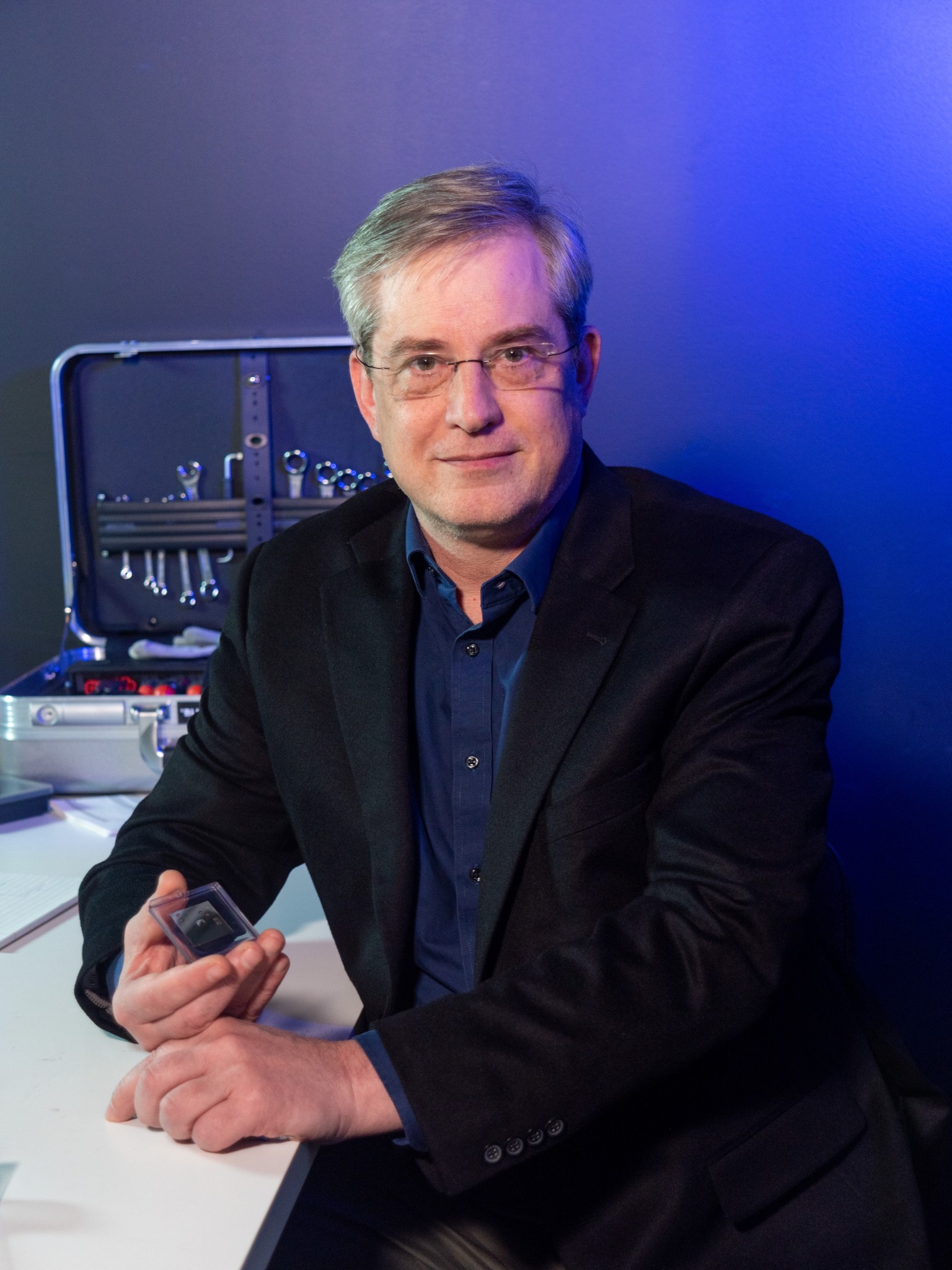 A man in black jacket and dark blue shirt sits at a desk, holding a small rectangle encased in clear covering. Behind him to his left, a toolbox sits open on the desk, and the wall is lit with a blue hue that fades to gray and black.