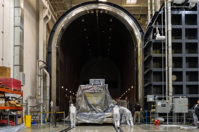 A wide shot of a silvery piece of space hardware about the size of a car sitting on a silver platform. Three techs in white head-to-toe suits are pushing it from behind through a very large, arched doorway that leads to a dark room lined with dim lights.