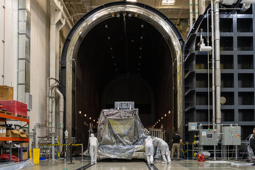 A wide shot of a silvery piece of space hardware about the size of a car sitting on a silver platform. Three techs in white head-to-toe suits are pushing it from behind through a very large, arched doorway that leads to a dark room lined with dim lights.