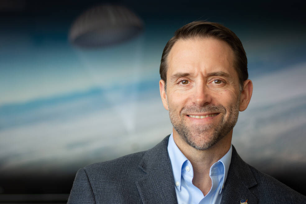 Mike Moreau, a man with dark brown hair and a short beard, smiles at the camera in a formal portrait in front of an illustration of the OSIRIS-REx capsule. He wears a blue woven suit with a light blue shirt and a NASA pin on his left lapel.