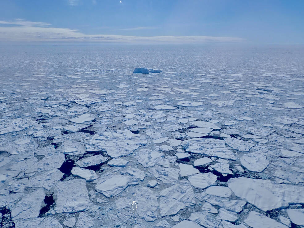 A flat expanse of broken white sea ice, jagged irregular polygons of flat white ice with spots of dark blue sea peeking through. The sea ice fades into the far distance where it meets the light blue sky. In the middle distance, a single blue iceberg rises above the sea ice.