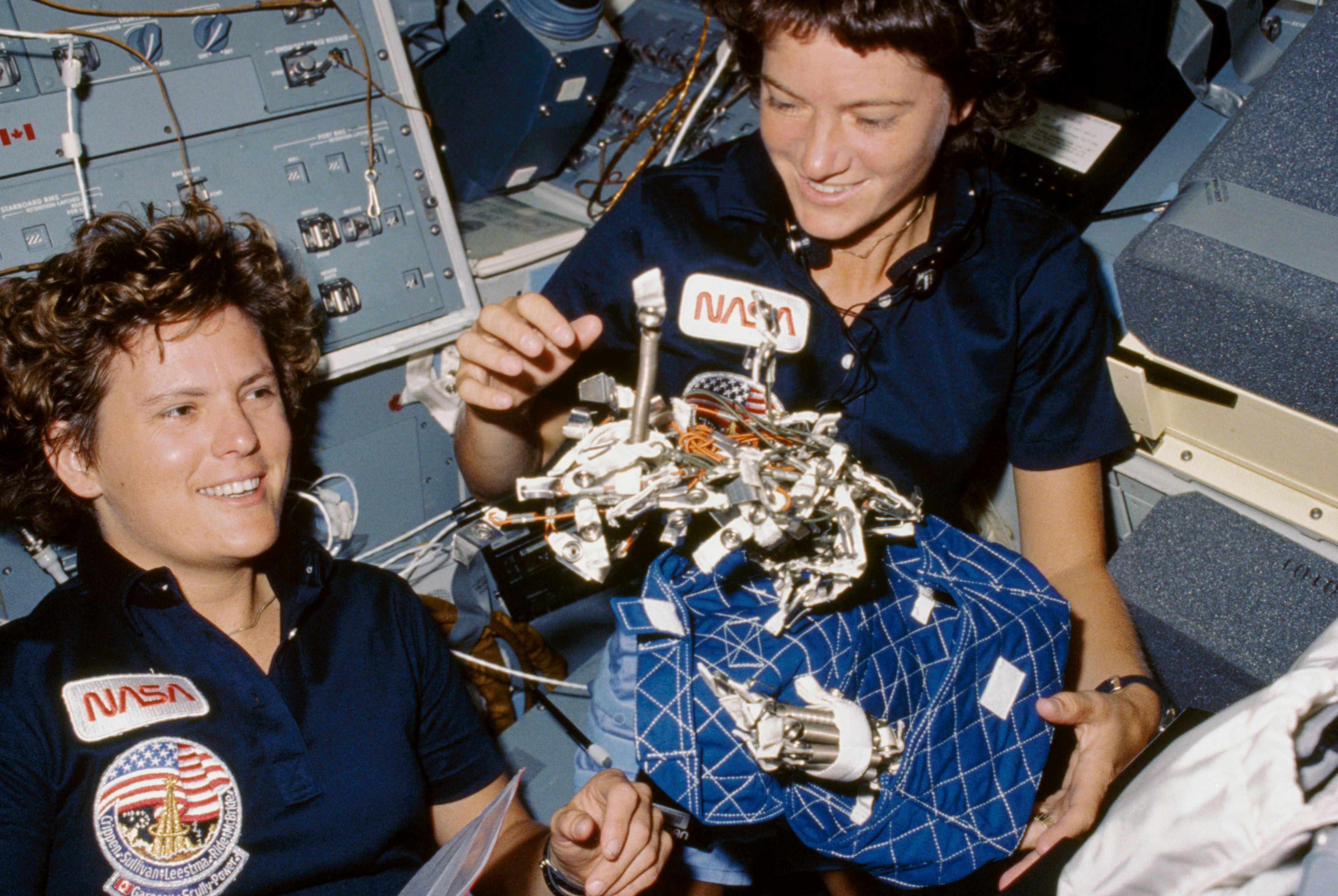 Astronauts Kathryn D. Sullivan, left, and Sally K. Ride display a "bag of worms". The "bag" is a sleep restraint and the majority of the "worms" ar...