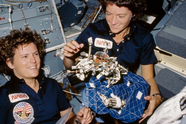 Astronauts Kathryn D. Sullivan, left, and Sally K. Ride display a "bag of worms". The "bag" is a sleep restraint and the majority of the "worms" are springs and clips used with the sleep restraint in its normal application. Clamps, a bungee cord and velcro strips are other recognizable items in the "bag".