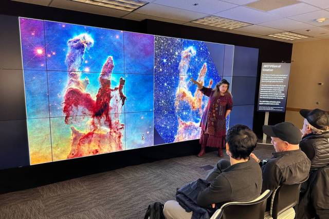 Michelle Thaller presents to a group of people, her hands across a large screen with a side-by-side of the Pillars of Creation taken by the James Webb Space Telescope and the Hubble Space Telescope.