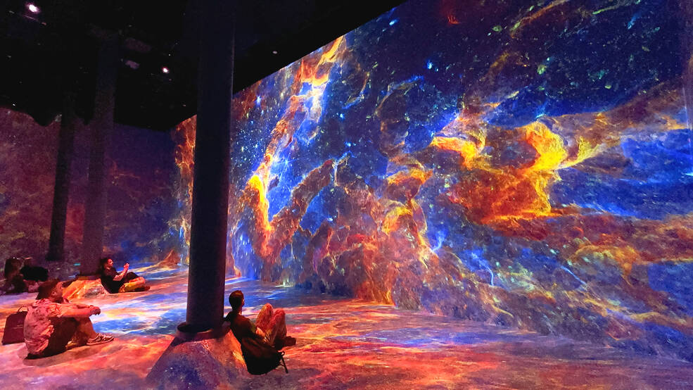 Attendees sit on the ground in a large art exhibit. The walls and floor are covered in projections inspired by Webb Telescope imagery of the Pillars of Creation.