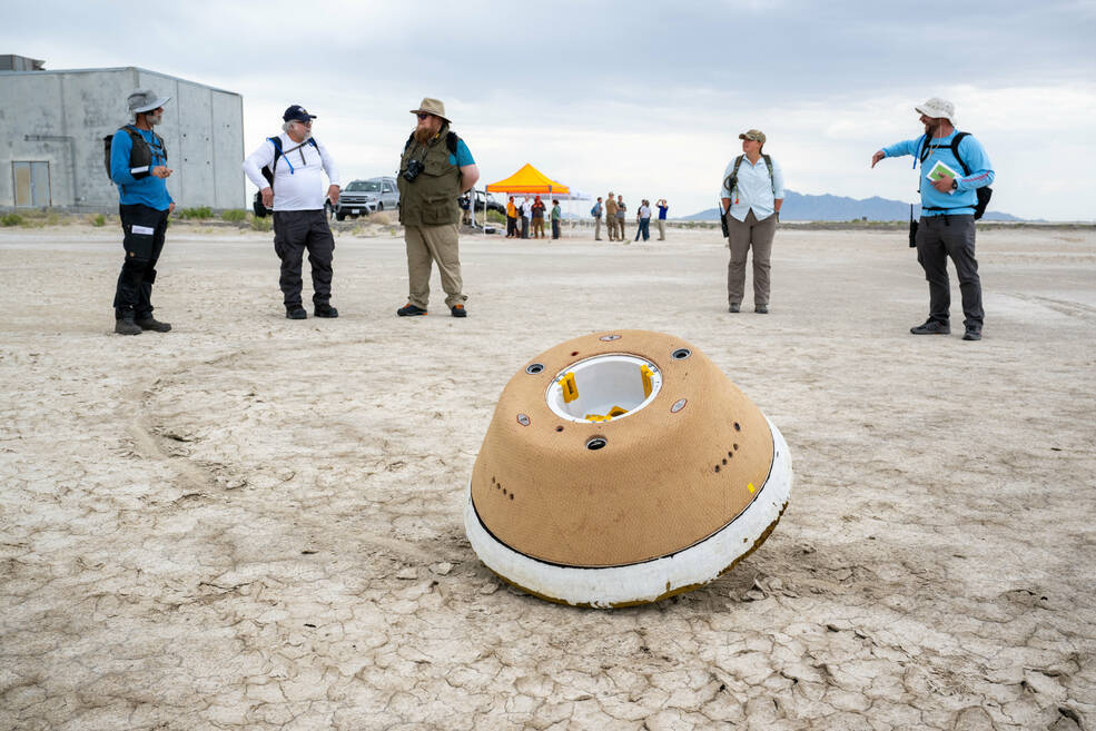 A stand-in for the OSIRIS-REx sample return capsule used for rehearsal of recovery operations sits on the cracked desert ground with a line of five people in the background.