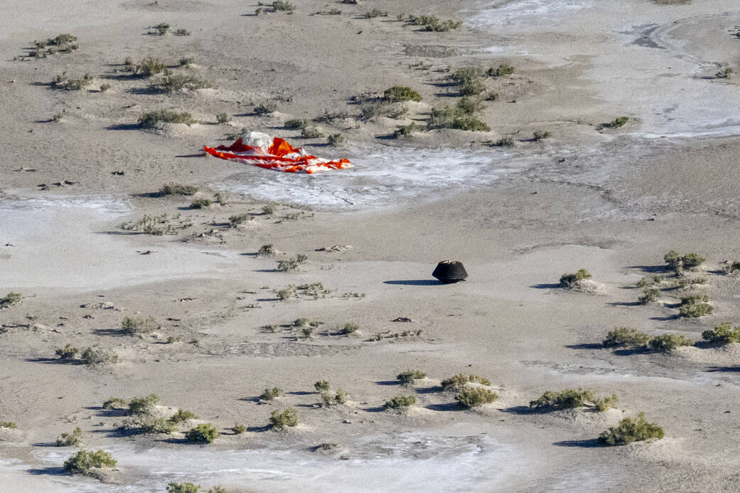 View from above of a desert landscape with brush. The red and white striped parachute that slowed the descent of the Sample Return Capsule is on the ground near the upper left and the black capsule is near the center of the image.