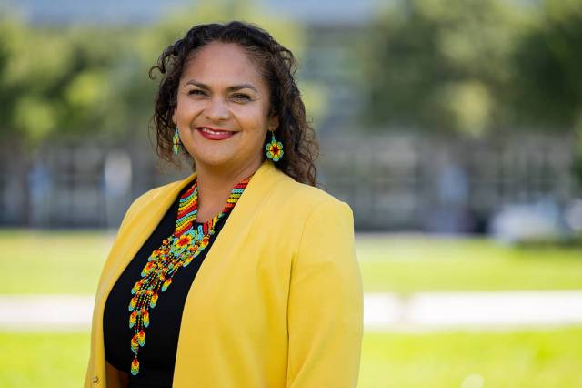 Carolina Rudisel smiles brightly as she looks at the camera. She is wearing a yellow jacket and black top, paired with a long rainbow-colored necklace and set of flower-shaped earrings, and her wavy brown hair frames her face. She is standing outside with leafy green trees and grass visible behind her.