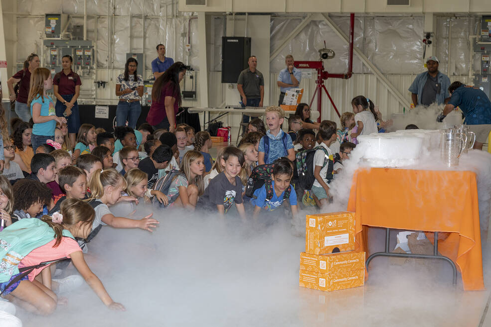 Group of young children are smiling and watching a fog/cloud like substance approach the ground.