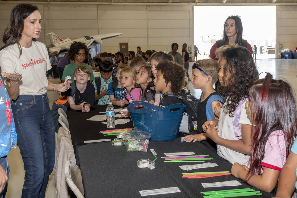 Group of young children watch as an adult with long black hair, wearing a white t-shirt and jeans points to a plane model.