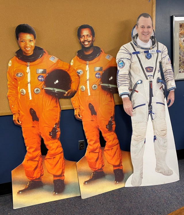 A photo of three life-sized cutouts of NASA astronauts. The two on the left are women wearing orange flight suits and holding helmets under their left arms. The one on the right is a man in a white flight suit. All of the astronauts have many patches and insignia on their uniforms.