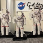A photo of three cutouts of white astronaut suits, with open circles where the helmets should be. The cutouts are designed for people of different sizes to stand behind the cutouts and look through the holes, so it looks as if they are "wearing" astronaut suits. The three cutouts stand in a lobby, in front of a wall sign with the NASA logo and "Goddard Space Flight Center".