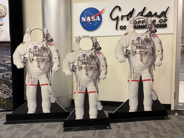 A photo of three cutouts of white astronaut suits, with open circles where the helmets should be. The cutouts are designed for people of different sizes to stand behind the cutouts and look through the holes, so it looks as if they are "wearing" astronaut suits. The three cutouts stand in a lobby, in front of a wall sign with the NASA logo and "Goddard Space Flight Center".