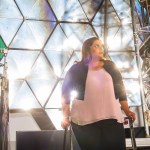 Abigail Rodriguez poses in front of the concentrator mirror in the Electric Propulsion and Power Laboratory at NASA’s Glenn Research Center. She is standing on a ladder and looks to her left. She is wearing a pink shirt, black cardigan, and black pants.