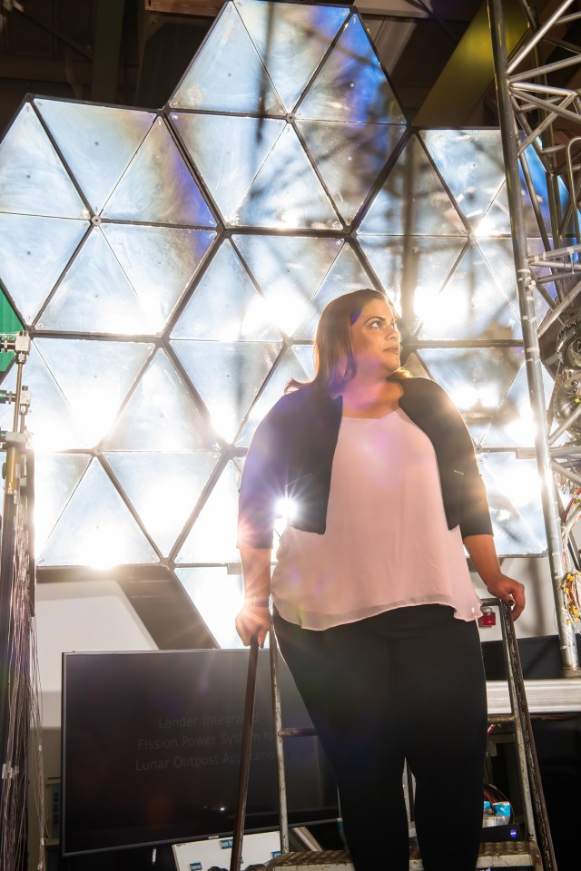 Abigail Rodriguez poses in front of the concentrator mirror in the Electric Propulsion and Power Laboratory at NASA’s Glenn Research Center. She is standing on a ladder and looks to her left. She is wearing a pink shirt, black cardigan, and black pants.