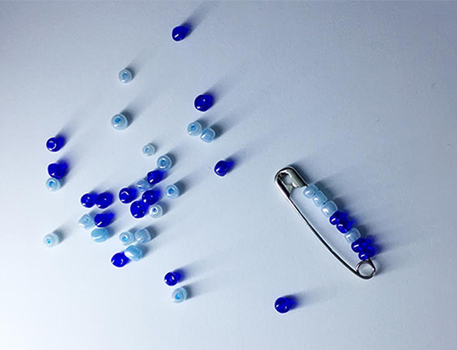 A safetypin with light blue and dark blue beads on it, surrounded by more light and dark blue beads scattered on the surface.