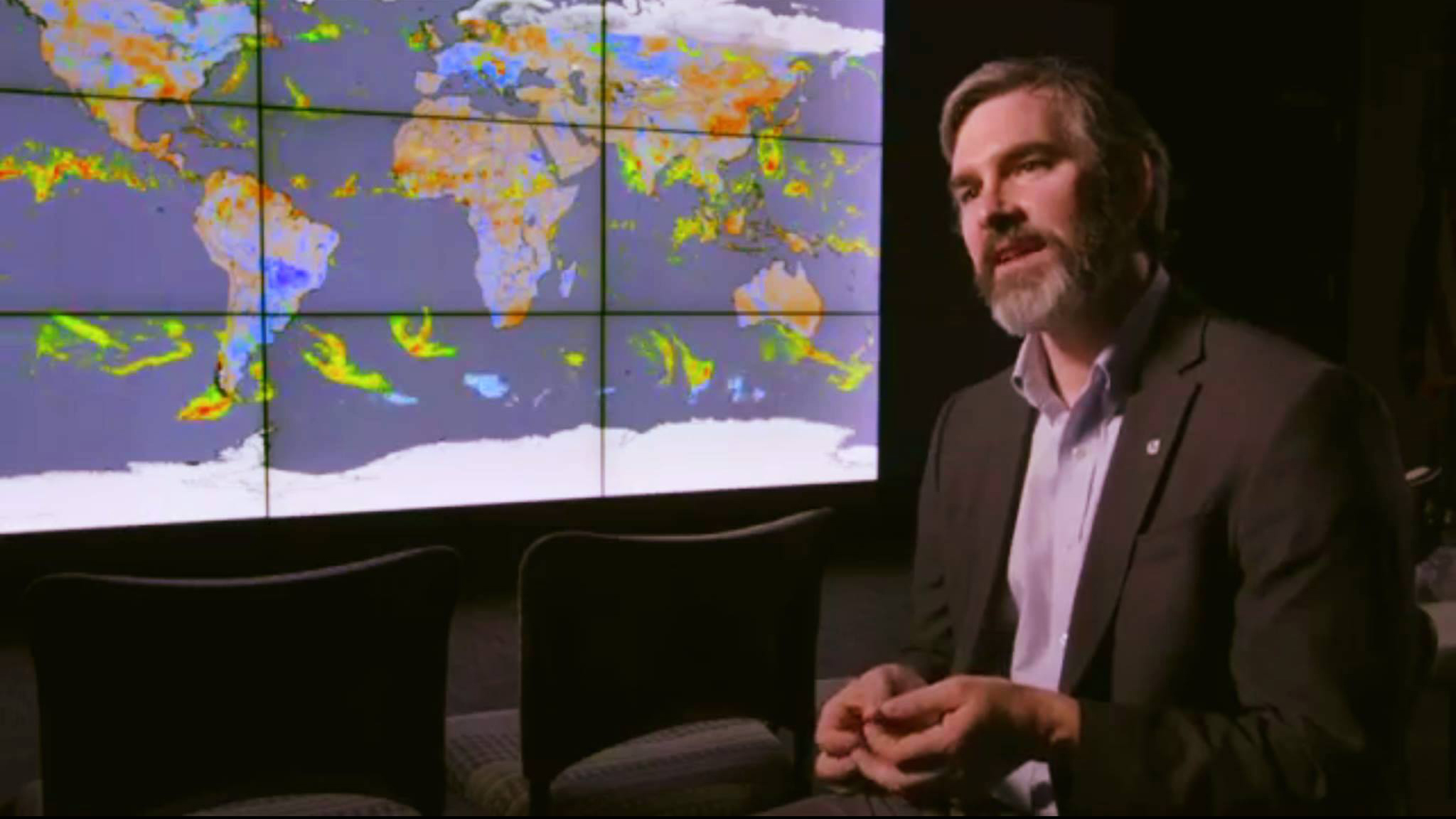man in suit with a bank of computer monitors projecting a map of Earth in the background
