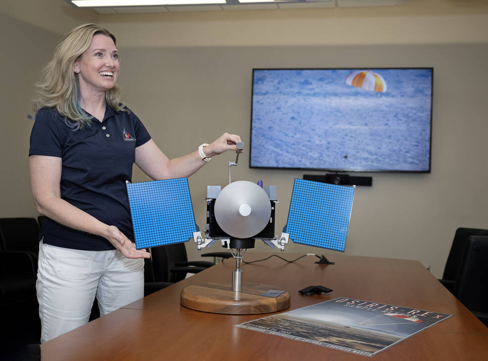 A blonde woman stands in front of a OSIRIS-REx model and smiles in a conference room.