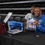 Retired NASA astronaut Leland Melvin smiles in his blue fight suit next to a young boy while holding a poster of The Color of Space he signed behind a table.