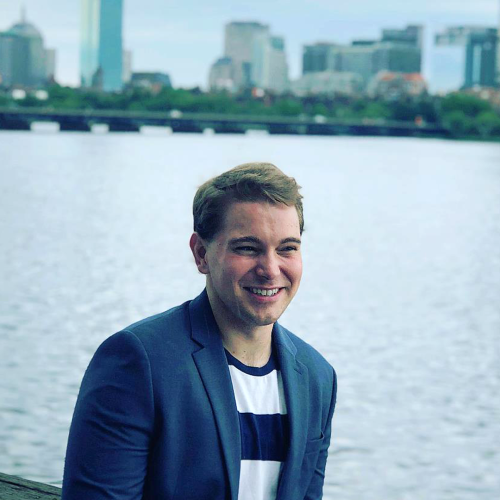 A smiling man with pale skin and short blonde hair is dressed in a blue suit and stands in front of the Charles River in Boston.