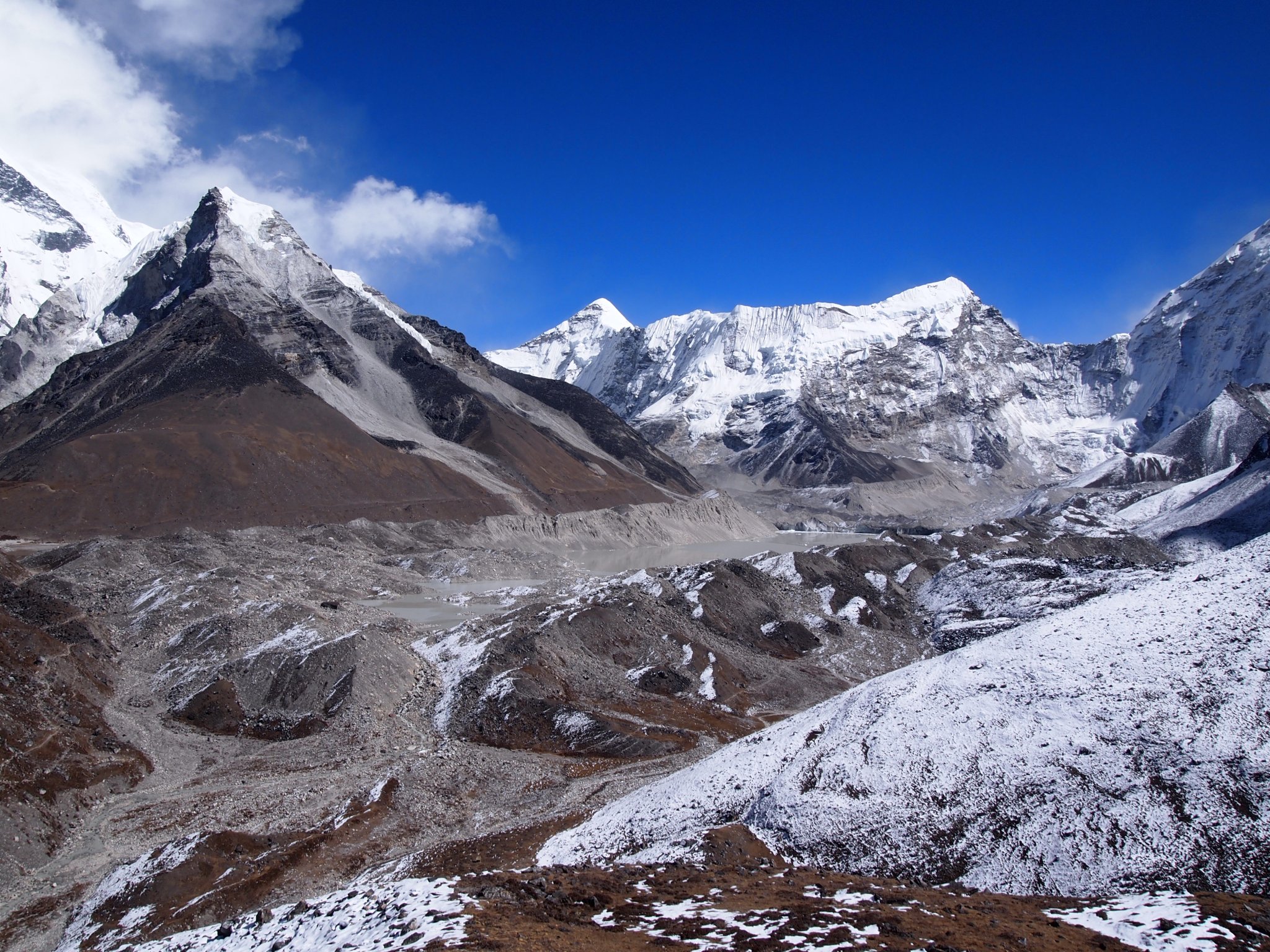 A photo of Imja Tsho, a lake made up of meltwater from Imja-Lhotse Shar Glacier in Eastern Nepal. One tall, sharp gray and brown mountain dominates the left side of the image, with gravelly lowlands partly covered with snow in the foreground. Peaks further back are covered with white snow. The sky is bright blue with a few puffy clouds above the largest mountain.