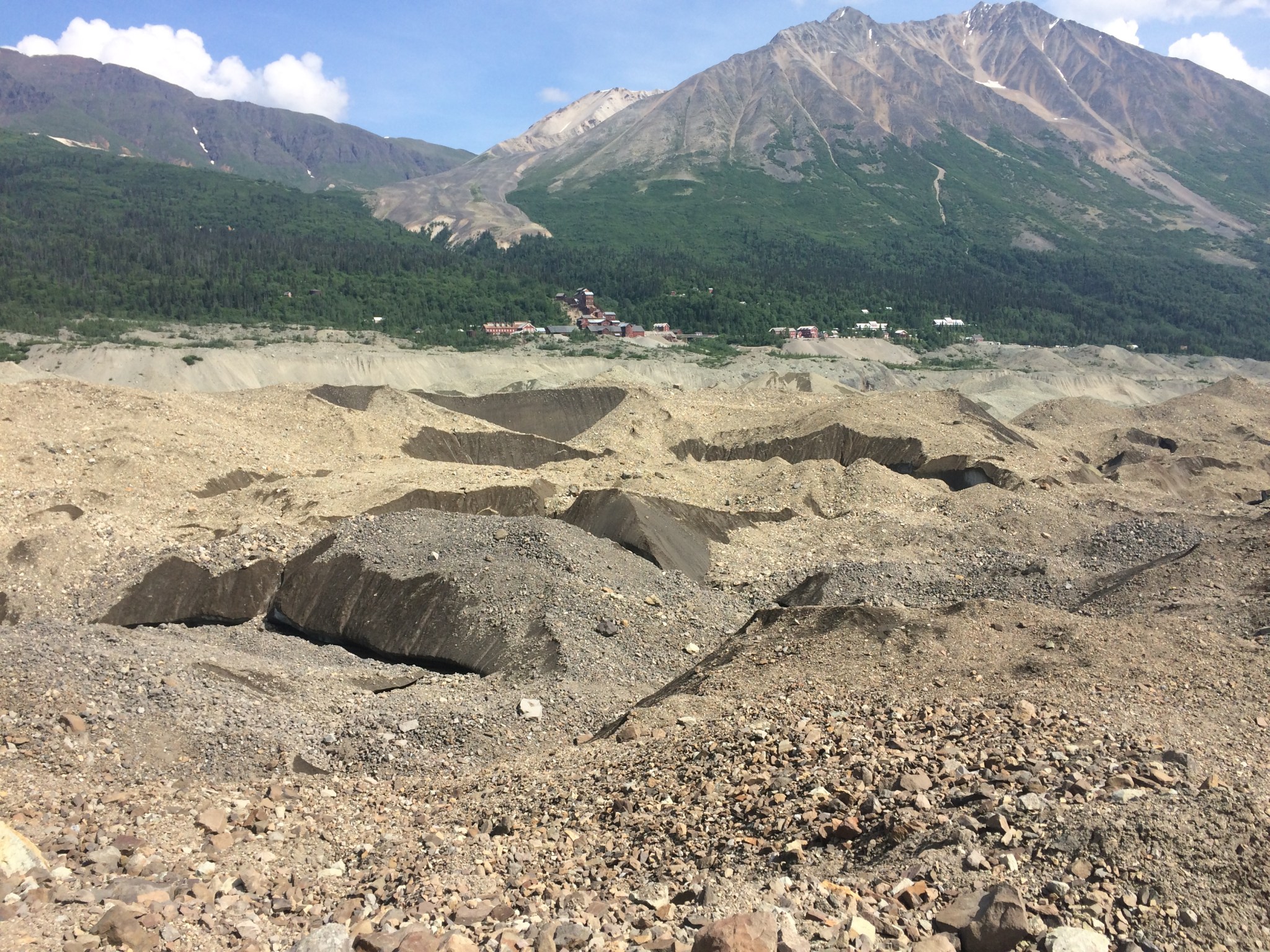 A photo of a glacier in Alaska, covered by tan and gray debris made up of soil, rocks, and ash. The glacier covers most of the foreground of the image - near the viewer, tiny rocks in many shades of brown and tan are visible. Further away, it looks more like sand, piled in peaks and piles with darker gray rocks showing through the soil. In the distance, the soil fades into dark green forests that spread up the sides of two tall gray-brown mountains. A small town made of red brick buildings is nestled between the two mountains. The sky is bright blue with fluffy white clouds.