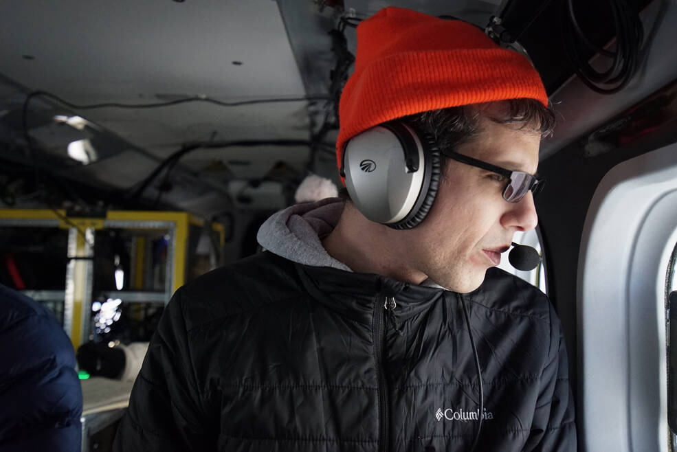 A man in a red knit cap looks out the window to the right in a darkened airplane. He wears headphones under the cap and a black parka. Behind him to the left, yellow frames hold racks of electronic equipment