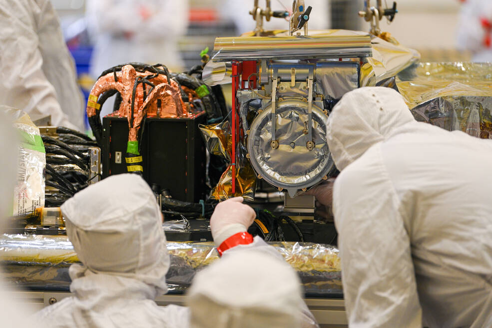 A technician in a white lab suit points to an instrument in front of them, with another technician in white lab suits looking closely at it. The instrument is silver and circular, surrounded by lots of wiring.