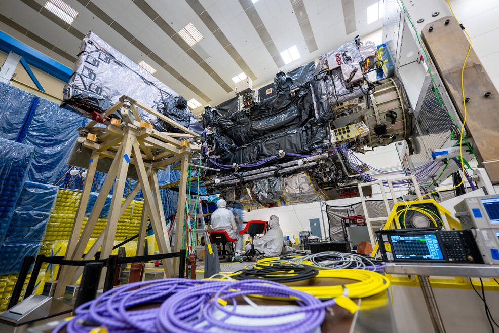 Technians in white lab suits work underneath the GOES-U satellite, which is mounted above them and covered in static bags.