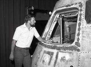 A black-and-white photo of a man in a polo shirt looking inside the Skylab 3 Apollo Command Module. One of the windows of the space capsule has been removed so visitors can view the interior. The exterior has burns and scuff marks.