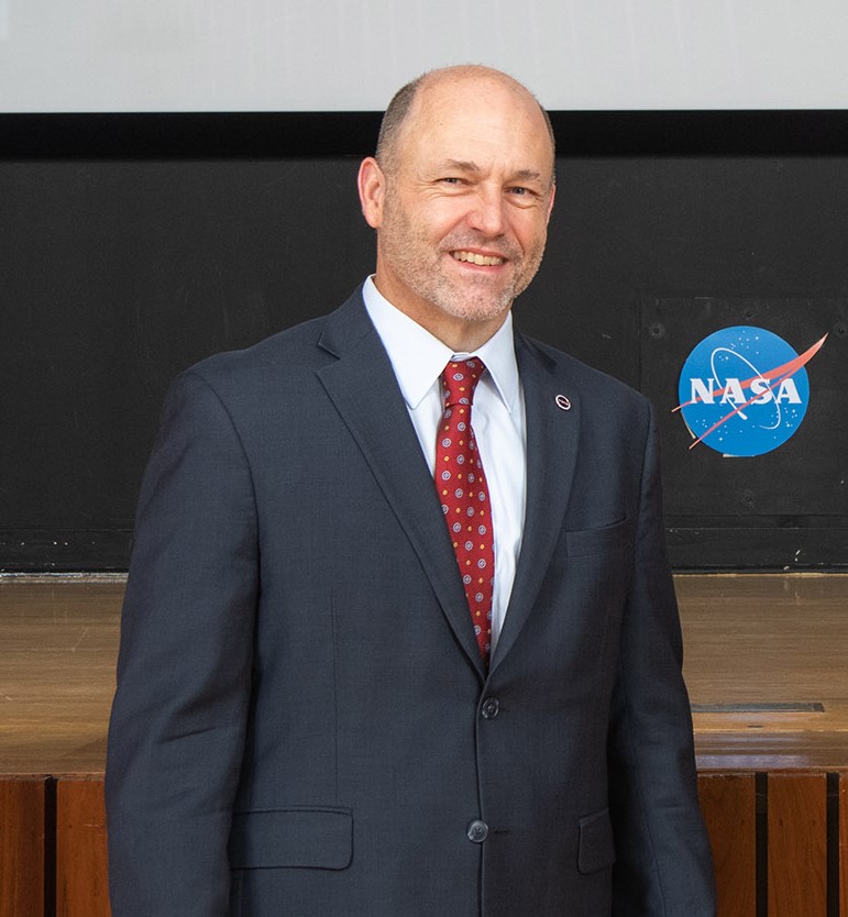 NASA Glenn center director Dr. James Kenyon poses during an Oath of Office ceremony. He is smiling and wearing a suit, A NASA meatball logo is seen in the background.