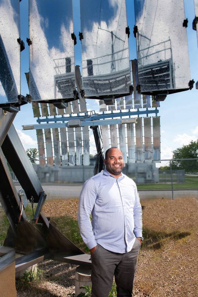Michael Pepen poses outside on NASA Glenn’s campus. He is standing next to a solar concentrator, a large metal structure with many reflective panels that extend above his head. He is smiling and wearing professional attire.